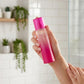 Scène : Photographie de style lifestyle. Une main féminine soignée tient délicatement le flacon "Simply Pink by Pink Sugar", s'apprêtant à l'utiliser après une douche. La main est positionnée naturellement. La lumière est douce et flatteuse.  Angle : Prise de vue à niveau, légèrement oblique.  Fond : Une salle de bain minimaliste floue, avec des carreaux de métro blancs et des plantes vertes. (gemini-3.1-flash-image-preview)