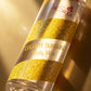 Angle : Gros plan macro serré avec un angle à $45^\circ$, orienté vers le centre du flacon.Scène : Focus absolu sur le flacon de "Creamy Sunshine" de l'image image_7cad21.jpg. Un rayon de soleil direct et chaud traverse l'image de biais, frappant de plein fouet les spirales jaunes pailletées. La texture granuleuse du glitter s'illumine en libérant des reflets holographiques dorés, orangés et argentés. L'inscription "Creamy Sunshine Body Mist" en lettres jaunes dorées est d'une netteté parfaite.Fond : Un arr