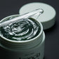 Angle : Gros plan macro très serré, focus sur la surface du gel ondulé.  Scène : Le pot d'Arencia de l'image image_c21e9d.jpg est ouvert, révélant la texture gélifiée et brillante du sérum vert forêt. Le couvercle mat céladon est posé en arrière-plan, légèrement flou. Une spatule en verre transparent est délicatement posée sur le rebord du pot, un fil de sérum brillant s'étirant depuis sa pointe.  Fond & Lumière : Arrière-plan sombre et minimaliste. Un éclairage rasant de studio fait miroiter les ondulation
