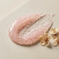Angle : Gros plan macro très serré, angle incliné à $35^\circ$ vers la texture.Scène : Focus extrême sur une noisette généreuse de gel-crème transparent/rose très clair (le peeling gel texturé de l'image) étalée au doigt sur une dalle de béton ciré crème mat. La texture révèle de minuscules granules de riz en suspension et une finition douce "gommage", prête à s'émulsionner. Quelques fleurs d'abricot séchées et texturées sont pressées à côté de la texture.Fond & Éclairage : Fond minimaliste crème mat. Éclai