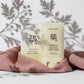 Une prise de vue frontale épurée et géométrique. Le stick solaire fermé et son packaging de l'image sont positionnés au centre Exact du cadre, debout. Leur base est délicatement enveloppée par un drapé de tissu de lin lourd couleur vieux rose poudré.  Détails : Fond monochrome blanc mat. Une lumière de studio directionnelle crue vient de biais de manière ciblée, projetant une ombre chinoise floue, poétique et parfaitement découpée de feuilles d'armoise et de baies sur le mur du fond et en superposition sur 