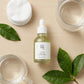 L'Angle : Vue de dessus, à plat (flat lay), mais organisée de manière organique.  Le Fond : Une table en bois clair. La bouteille de sérum est le point focal.  Atmosphère : Apaisant, organisé.  Le Prompt :  Organically arranged flat-lay product photography of a Beauty of Joseon serum bottle (image_1.png) as the central element. It is surrounded by other elements of a calming routine: a stack of clean white cotton rounds, a simple clear glass water pitcher, and a couple of perfect, large fresh green tea leav