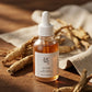 L'Angle : Vue en contre-plongée légère (low angle) pour donner de l'importance au produit.  Le Fond : Une table en bois de cèdre foncé, entourée de racines de ginseng séchées et d'un tissu en lin brut beige.  Le Prompt : > Commercial product photography of a Beauty of Joseon amber glass serum bottle. The bottle is standing on a dark, rustic wooden table. In the blurred background, raw ginseng roots and a piece of textured beige linen fabric. Warm, soft morning light coming from the side, casting gentle shad