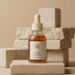 L'Angle : Vue de face (eye-level shot), parfaitement centrée.  Le Fond : Des blocs de pierre calcaire ou de travertin beige beige clair empilés de manière asymétrique, sur un fond monochrome sable.  Le Prompt : > Minimalist e-commerce studio shot of an amber skincare dropper bottle. The product is placed on a smooth beige travertine stone block. Clean, monochromatic sand-colored background. Soft, diffused overhead lighting, clean studio lighting, hyper-realistic glass texture, premium cosmetics branding loo