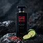 Commercial product photography of a single large 650ml opaque black shower gel bottle with a metallic red label reading "ROSE INDOMPTABLE - GRIS MONTAIGNE PARIS". The bottle is standing alone on a wet, dark volcanic basalt rock surface. In the foreground, fresh slices of green bergamot and scattered crushed pink peppercorns are artfully arranged. Fine water droplets are misting in the air, catching a dramatic side light. Dark, atmospheric, high contrast, clean product focus, e-commerce catalog quality. (gem