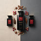 An elegant top-down flat-lay product photograph for an e-commerce website. Three black and glass fragrance bottles from the "Rose Indomptable" collection are arranged perfectly parallel on a modern split-background: one half is natural beige textured linen, the other half is smooth matte charcoal concrete. Interspersed neatly between the bottles are raw natural ingredients: dried rosebuds, small pieces of precious dark wood, and delicate spices. Clean, symmetrical composition, soft even daylight, overhead s