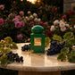 Un plan large d'un flacon de image_0.png posé sur une table en travertin poli, sur une terrasse donnant sur un jardin de roses anciennes à la tombée de la nuit. La lumière est chaude et dorée (golden hour). Autour de image_0.png, des grappes de raisin de Corinthe et des feuilles de géranium créent un cadre organique. La plaque dorée de image_0.png reflète l'heure dorée. Style : Photographie éditoriale de luxe, sensuelle. (gemini-3.1-flash-image-preview)