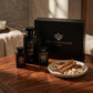Un plan large à 45 degrés du coffret et des trois flacons de image_2210be.jpg exposés sur une table en bois d'acajou précieux et poli. À côté des bouteilles, une coupelle en céramique artisanale contient des bâtons de cannelle et de l'encens brut. Une lumière douce et rasante traverse la pièce, installant une atmosphère chaleureuse et boisée de spa privé et de soin quotidien de luxe. Style : Campagne éditoriale luxueuse. (gemini-3.1-flash-image-preview)