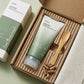 L'arrière-plan est le carton d'emballage du produit (que nous pouvons supposer être similaire en style, blanc avec du texte vert). Le tube Anua est disposé à l'intérieur de l'insert en carton ondulé texturé et écologique. À côté, on voit une petite cuillère d'application en bambou et quelques capsules d'un supplément de quercétine. Cela renforce l'aspect naturel et le lien avec l'ingrédient principal.  Angle : Vue de dessus, légèrement inclinée pour donner de la profondeur à la boîte.  Fonds : Carton d'emba