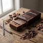 Photographie de style food styling mettant en scène le flacon de parfum Arabiyat Sugar "Chocolate Ganache" (comme vu dans image_0.png) glissant doucement hors de sa boîte ouverte, posée à plat sur une table en bois rustique patiné par le temps. Le flacon, avec sa base texturée "biscuit" et ses coulures de chocolat mates, est à moitié sorti, laissant apparaître l'ouverture du carton imprimé "carré de chocolat". Tout autour de la boîte et du flacon, sont dispersés de manière naturelle des ingrédients bruts : 