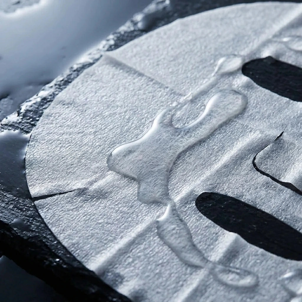 Angle : Gros plan macro très serré, angle à $45^\circ$.Scène : Le masque en tissu blanc de l'image image_cdfb9f.jpg, sorti de son sachet, est délicatement déplié et posé à plat sur une surface sombre. La finesse extrême du tissu est mise en avant : il est si gorgé de sérum qu'il devient presque transparent par endroits. De riches gouttes de sérum limpide et visqueux coulent le long de ses fibres texturées.Décor & Lumière : Arrière-plan flou en ardoise noire humide. Une lumière de studio rasante et froide fa