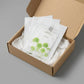 Scène : Trois sachets de masques de l'image image_cdfb9f.jpg sont présentés empilés de manière légèrement décalée à l'intérieur d'un coffret de livraison premium en carton kraft beige ouvert. Les feuilles vertes du sachet du dessus apportent une touche de couleur éclatante au milieu de papiers de soie froissés blanc laiteux.  Fond & Lumière : Surface de studio mate et contemporaine grise. Éclairage uniforme et très lumineux, mettant en valeur l'aspect e-commerce premium. (gemini-3-pro-image-preview)