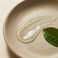 Angle : Gros plan macro très serré, angle incliné à $35^\circ$ braqué sur la texture.Scène : Une trace généreuse et brillante de sérum liquide et aqueux (la texture de l'image image_cf5cde.png) est étalée délicatement sur une assiette en céramique artisanale beige mat au fini rugueux. La formule est parfaitement transparente, fluide et emprisonne deux ou trois micro-bulles d'air. Une feuille de thé vert fraîche est posée juste à côté de la texture.Décor & Éclairage : Fond épuré ton-sur-ton crème. Éclairage 