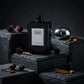 Concept de shooting : Utiliser des plaques de pierres naturelles de différentes hauteurs pour créer du relief.  Le Prompt à copier :  Une photographie de nature morte e-commerce. La boîte noire "BLACK EMPIRE" et son flacon laqué assorti sont mis en scène sur des blocs de tailles différentes en ardoise noire brute et feuilletée, créant un jeu de marches géométriques. Quelques boutons de rose séchés sombres et des éclats de bois de oud sont parsemés sur les pierres. La plaque argentée du parfum accroche la lu