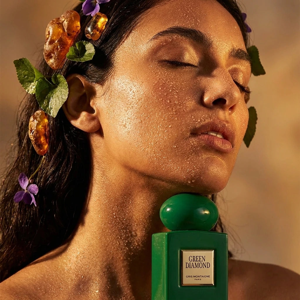 Un gros plan d'un mannequin (homme ou femme), sa peau légèrement couverte d'une rosée fine. Son cou est visible, avec le flacon de image_0.png reposant à sa base, comme un bijou. Ses yeux sont fermés, expression d'extase. Ses cheveux sont parsemés de petits cristaux d'ambre brut et de feuilles de violette. La lumière est chaude et rasante, accentuant les textures. Style : Photographie de beauté conceptuelle, texture de peau riche. (gemini-3-pro-image-preview)
