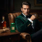 Un plan moyen d'un mannequin homme au visage sculpté, assis dans un fauteuil en cuir vieilli. Le flacon de image_0.png est posé sur une table d'appoint à côté de lui, avec une bouteille de cognac ambrée et un fagot de racines de vétiver. Il porte un smoking vert émeraude foncé. Son regard est direct. La plaque dorée de image_0.png s'aligne avec ses boutons de manchette. L'éclairage est sombre, contrasté. Style : Photographie de portrait d'affaires de luxe. (gemini-3-pro-image-preview)