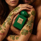 Un plan en plongée rapproché de deux personnes (une femme et un homme) dont les bras s'entrelacent autour du flacon de image_0.png. Leurs bras sont peints de motifs organiques de mousse de chêne et de feuilles de géranium, se fondant l'un dans l'autre. Le flacon de image_0.png est le cœur de cette étreinte, sa plaque dorée reflétant leurs peaux. La lumière est tamisée, chaude et intime. Style : Photographie d'art corporelle, composition tactile. (gemini-3-pro-image-preview)