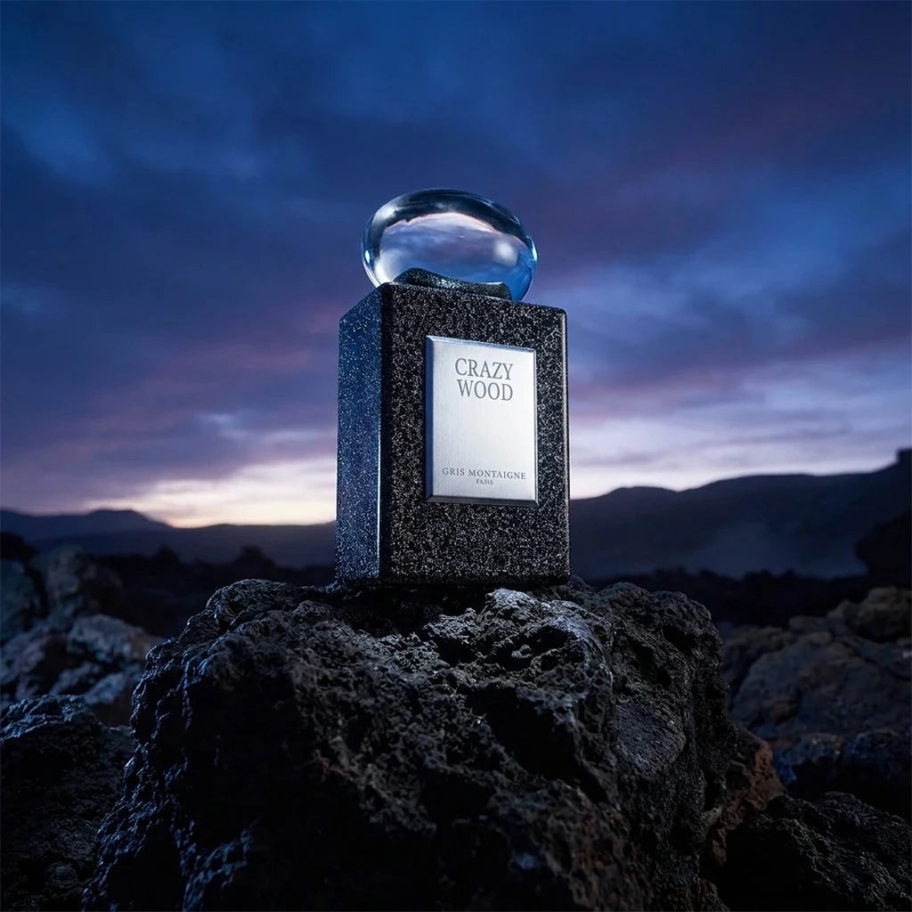 Un plan large en contre-plongée (vue basse) plaçant le flacon de image_5.png sur un piédestal de roche volcanique brute. Le ciel en arrière-plan est sombre, juste avant l’aube, apportant une lueur bleu nuit froide. La plaque argentée 'CRAZY WOOD' au centre de image_5.png attrape la seule source de lumière studio blanche, la rendant parfaitement lisible et contrastée face au bloc rocheux. Style : Photographie de paysage conceptuelle pour parfum. (gemini-3-pro-image-preview)