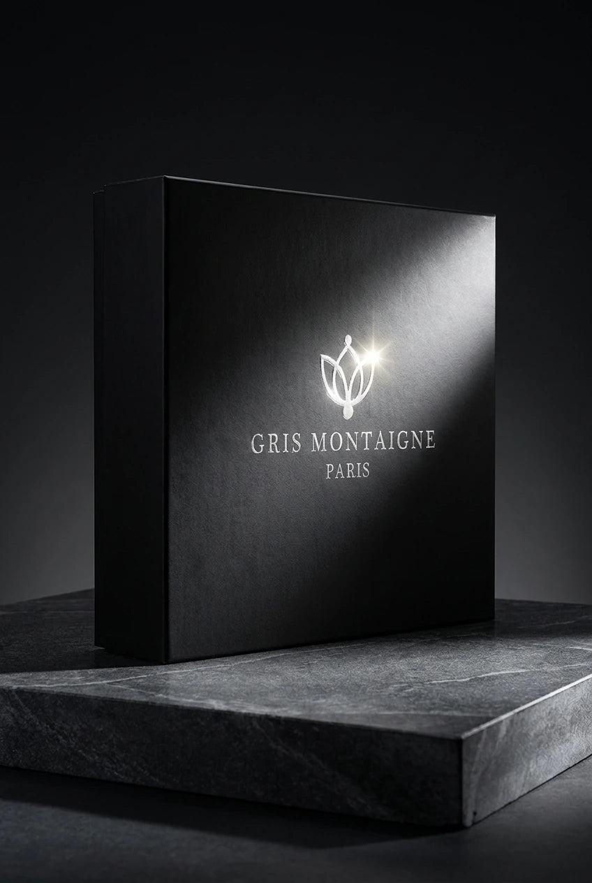 Un plan moyen net et rigoureusement frontal du grand coffret cadeau noir fermé de image_2210be.jpg posé à la verticale sur une dalle de grès poli gris anthracite texturé. La lumière de studio est directionnelle, venant frapper de biais le logo argenté central de image_2210be.jpg pour créer un éclat brillant ultra-réaliste. L'arrière-plan est sombre et épuré. Style : Photographie publicitaire de haute parfumerie, clair-obscur sophistiqué. (gemini-3-pro-image-preview)