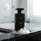 Un plan moyen de profil montrant uniquement l'imposant flacon de gel douche noir de image_2210be.jpg posé sur le rebord en marbre noir d'une douche moderne. Une texture riche, vaporeuse et aérienne de mousse blanche onctueuse est délicatement déposée à sa base, contrastant avec l'opacité sombre de la bouteille. La lumière naturelle filtre à travers une vitre sablée. Style : Photographie lifestyle haut de gamme. (gemini-3-pro-image-preview)