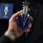 Un plan serré et intimiste centré sur les mains d'un mannequin homme au poignet orné d'une montre en acier brossé. Ses doigts tiennent fermement et avec assurance le corps sculptural en verre bleu et chrome du flacon de image_2d17cf.jpg, extrait de son coffret visible en arrière-plan flou. Le fond de l'image montre la texture noble d'une veste de costume noire sur mesure. Style : Campagne publicitaire éditoriale de mode et de parfum masculin. (gemini-3-pro-image-preview)