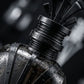 Un plan macro très serré concentré sur le col métallique marqué 'GRIS MONTAIGNE' et la couronne impériale noire de image_2de6bf.jpg. De fines micro-gouttelettes d'eau pure perlent de manière réaliste sur la surface externe du verre fumé transparent, évoquant une fraîcheur divine. L'arrière-plan présente un flou artistique (bokeh) très sombre composé de douces lueurs argentées et noires. Style : Photographie de produit sensorielle et haut de gamme. (gemini-3-pro-image-preview)
