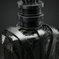 Un plan macro très rapproché concentré sur le col métallique noir gravé 'GRIS MONTAIGNE' et les structures géométriques de image_2ee9ff.jpg. De minuscules micro-gouttes de condensation fraîche perlent de manière ultra-réaliste sur la surface lisse en verre fumé, contrastant avec le fini mat des arêtes en métal noir de image_2ee9ff.jpg. L'arrière-plan présente un flou artistique (bokeh) sombre aux nuances gris anthracite. Style : Photographie de produit hautement sensorielle. (gemini-3-pro-image-preview)