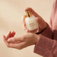 Un plan serré concentré sur les mains délicates d'un mannequin femme à la peau douce, vêtue d'un pull en maille de cachemire vieux rose texturé. Ses doigts aux ongles soignés tiennent avec grâce le flacon transparent de image_30acb9.jpg, s'apprêtant à appliquer une goutte de ce musc crémeux sur le creux de son poignet. L'arrière-plan est plongé dans un flou beige ultra-lumineux. Style : Publicité de beauté intime et réconfortante. (gemini-3-pro-image-preview)