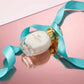Un plan moyen en plongée à 60 degrés du flacon de image_3aa887.jpg couché délicatement sur une surface miroir rose laquée parfaitement polie. Le miroir crée une réflexion symétrique, nette et impeccable de la bouteille ovale. Un long ruban de soie de couleur bleu turquoise ondule gracieusement autour du bouchon métallique doré de image_3aa887.jpg. Style : Composition artistique d'art contemporain. (gemini-3-pro-image-preview)