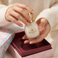 Un plan serré concentré sur les mains délicates d'un mannequin femme à la peau douce, vêtue d'un pull en maille de cachemire blanc texturé. Ses doigts aux ongles impeccables retirent délicatement le flacon transparent de image_3aa887.jpg de son compartiment intérieur bordeaux/rose. L'arrière-plan de l'écrin et ses frises turquoise de image_3aa887.jpg apparaissent dans un flou poétique et lumineux. Style : Publicité de beauté intime et réconfortante. (gemini-3-pro-image-preview)