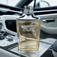 Une photographie publicitaire au format carré 1200x1200px. Le flacon de parfum "Bentley Infinite" de image_8532e2.jpg est photographié de face, parfaitement centré et d'une netteté absolue. Il repose sur la console centrale en fibre de carbone et aluminium brossé d'un coupé de sport moderne. L'arrière-plan présente l'intérieur minimaliste et technologique du véhicule avec des éclairages LED linéaires blancs et froids. La lumière du jour se reflète proprement sur les épaules chromées et met en relief l'emblè