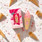 Une vue de dessus (flat-lay) sophistiquée sur un format carré de 1200x1200px. Le parfum et sa boîte d'image_943e3f.jpg sont disposés élégamment au centre d'un marbre blanc immaculé. Ils sont entourés de véritables mini-cornets de glace vides texturés, de paillettes comestibles roses éparpillées et de délicates touches de crème de lait blanche. L'ensemble est baigné d'une lumière estivale lumineuse, fraîche et haut de gamme. (gpt-image-2)
